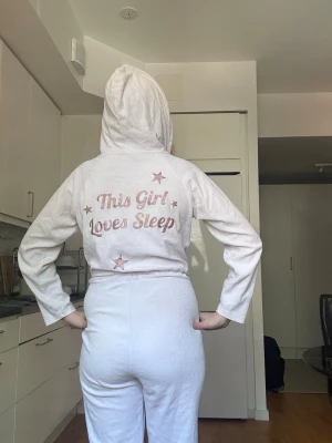 Ljusrosa onesie med huva och text - Mysig ljusrosa onesie med huva och dragkedja framtill. Resår i midjan med snörning för skön passform. På ryggen står det 'This Girl Loves Sleep' i glittrig rosa text och stjärnor. Perfekt för chill dagar hemma.