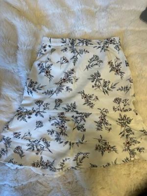 Vit blommig kjol med volang - Supersöt vit kjol med blommigt mönster och volangkant. Kjolen har en lätt utsvängd form!