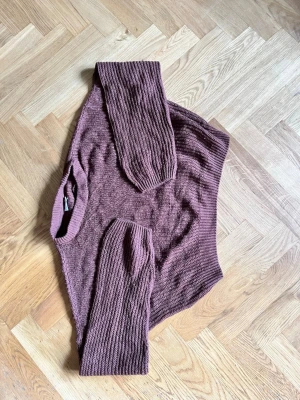 Brun stickad tröja med ribbade ärmar - Mysig brun stickad tröja med bred ribbstickning och rund hals. Tröjan har långa, löst sittande ärmar och en avslappnad passform. Perfekt för lager på lager och enkel att matcha med olika stilar.