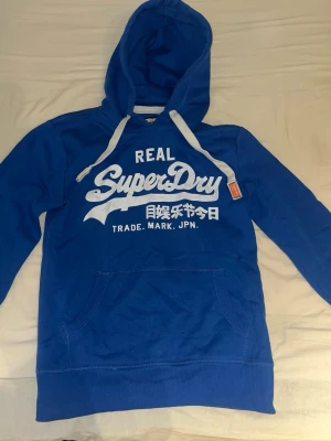 Blå Superdry hoodie med tryck - Snygg blå hoodie från Superdry med vit logotyp och japanska tecken på bröstet. Hoodien har en huva med vita snören och en praktisk magficka. Perfekt för dig som gillar streetwear och vill ha något bekvämt och stilrent.
