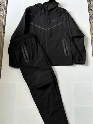 Nike tracksuit  - Snyggt svart träningsset från Nike med jacka och byxor. Jackan har huva, hel dragkedja och två fickor med dragkedja. Byxorna har resår i midjan och dragkedjeförsedda fickor. Setet har diskreta reflexdetaljer och är gjort i ett lätt, sportigt material.