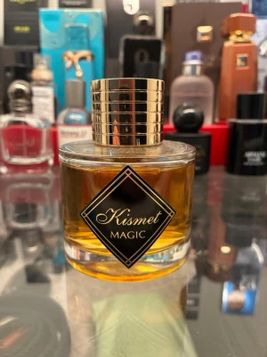 Kismet Magic Eau de Parfum - Kismet Magic Eau de Parfum med elegant rund glasflaska och guldig randig kork. Parfymen har en bärnstensfärgad vätska och en stilren svart etikett med guldiga detaljer. Perfekt för dig som gillar lyxiga och unika dofter.