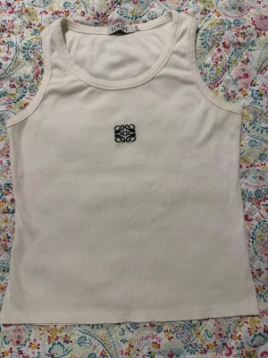 Vit ribbad linnetopp från Loewe - Säljer en vit ribbad linnetopp från Loewe med broderad svart logga på bröstet. Toppen är ärmlös och har en klassisk rund halsringning. Perfekt för dig som gillar stilrena och trendiga basplagg.