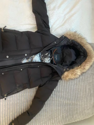 Svart vinterjacka med äkta Pälsfrån Hollies - Snygg svart vinterjacka från Hollies med avtagbar huva och äkta päls  runt kanten. Jackan har två dragkedjeförsedda fickor framtill och stängs med både dragkedja och knappar. Perfekt för kalla vinterdagar och riktigt varm. Ord: 5000 men säljs för 1100