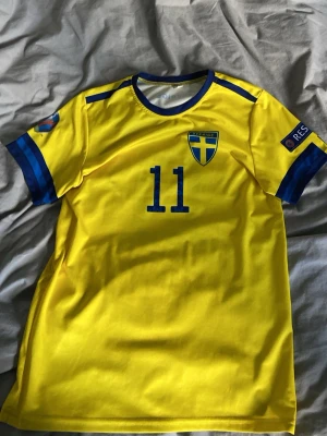 Sverige fotbollströja Isak #11 - Gul Sverige fotbollströja  med blå detaljer, nummer 11 och namnet Isak på ryggen. Tröjan har rund hals, blå ränder på ärmarna och Sveriges landslagsemblem på bröstet. Bara använd när Sverige spelat matcher
