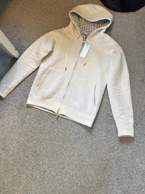 Dior zip - Snygg beige hoodie från Dior med dragkedja framtill och två stora fickor. Huvan har ett mönstrat innerfoder och dragsko med metalländar. Diskret Dior-logga broderad på fickan. Tillverkad i mjukt material som känns lyxigt och bekvämt.