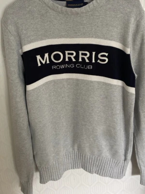 Morris tröja - Snygg grå stickad tröja från Morris Rowing Club med mörkblå och vit rand över bröstet och broderad logga. Tröjan har rund halsringning, långa ärmar och ribbade muddar. Perfekt för en sportig och avslappnad look. Storlek  L men passar som M.