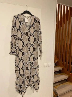 Mönstrad långklänning med v-ringning - Säljer en långklänning med svart och beige ormmönster. Klänningen har v-ringning, trekvartsärm och asymmetrisk volangkant nedtill. Materialet känns lätt och luftigt, perfekt för dig som gillar statementplagg.