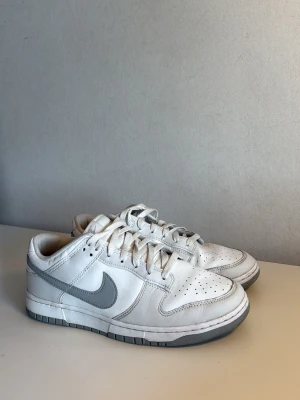 Nike Dunk Low vita och grå sneakers - Nike Dunk Low sneakers i vitt och ljusgrått läder. Säljs pga inte använder längre. Skorna är använda en gång för länge sen. Orginalkarton och kvitto medföljer ej tyvärr.