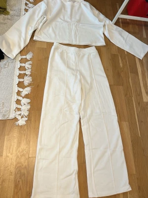 Vitt loungewear-set med vida byxor - Mysigt vitt loungewear-set till sommaren med långärmad, croppad tröja och matchande vida byxor med resår i midjan. Setet är tillverkat i mjukt tyg, perfekt för chill dagar hemma. Enkel och stilren design som känns både modern och bekväm. Oanvänd