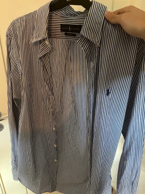 Randig skjorta från Ralph Lauren - Säljer en klassisk blåvit randig skjorta från Ralph Lauren i slim fit-modell. Skjortan har långa ärmar, knappar framtill och den ikoniska broderade loggan på bröstet. Perfekt för dig som gillar stilrena och tidlösa plagg.