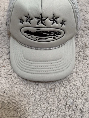 Grå meshkeps med broderade stjärnor - Snygg grå keps med mesh på baksidan och justerbar snapback. Framtill finns broderade svarta stjärnor och ett motiv, samt broderade detaljer även på sidan. Perfekt för dig som gillar streetwear och vill ha en unik accessoar.