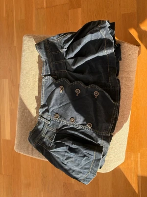 Vintage mörk jeanskjol  - Säljer en mörkblå jeanskjol med dekorativa knappar framtill och vågig kant. Kjolen har markerade sömmar och en cool, lite puffig form. .