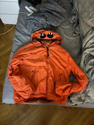 Orange vindjacka från C.P. Company - Snygg orange vindjacka från C.P. Company med ikoniska glasögon i huvan. Jackan har dragkedja framtill, två framfickor med knapp och justerbar huva. Tillverkad i lätt och slitstarkt material, perfekt för blåsiga dagar. STORLEK L, ÄKTA, BUDGIVNING. Aldrig använd då den var för stor och fick den som present, varit ihopvikt i låda