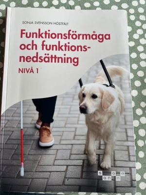 Funktionsförmåga och funktionsnedsättning - Kursbok med titeln 'Funktionsförmåga och funktionsnedsättning Nivå 1' av Sonja Svensson Höstfält. Omslaget är vitt med röd text och visar en ledarhund och en person med vit käpp. Perfekt för dig som pluggar vård och omsorg.