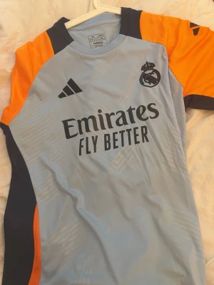 Real Madrid träningströja Adidas XL - Snygg Real Madrid träningströja från Adidas i ljusblått med orange och mörkblå detaljer. Tröjan har klubbmärke och Adidas-logga på bröstet samt 'Emirates Fly Better' tryckt framtill. Tillverkad i lätt och ventilerande material, perfekt för träning.
