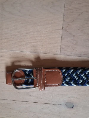 Flätat blått bälte med läderdetaljer - Snyggt flätat bälte i blått, vitt och grått med bruna läderdetaljer och silvrigt spänne. Perfekt accessoar för att lyfta din outfit med en sportig och stilren touch. Bältet har klassisk design och passar till både jeans och chinos.