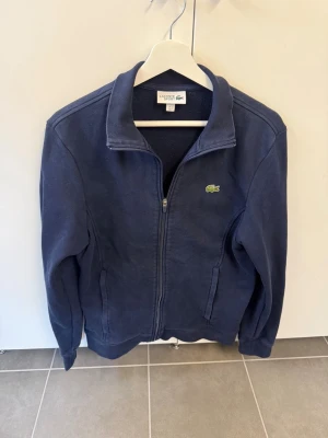 Marinblå fleecejacka från Lacoste, stl M - Säljer en marinblå fleecejacka från Lacoste Sport med dragkedja framtill och två fickor. Klassisk krokodillogga på bröstet och hög krage. Perfekt för dig som gillar sportig och stilren look.