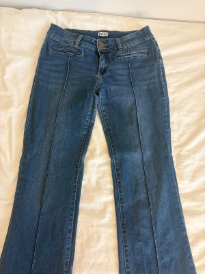 Blå bootcut jeans från Madlady - Säljer ett par blå bootcut jeans från Madlady med dubbla knappar i midjan och dekorativa sömmar framtill. Jeansen har klassisk femficksdesign och är tillverkade i denim med lätt stretch. Perfekt för dig som gillar en retrostil och vill ha bekväma jeans.