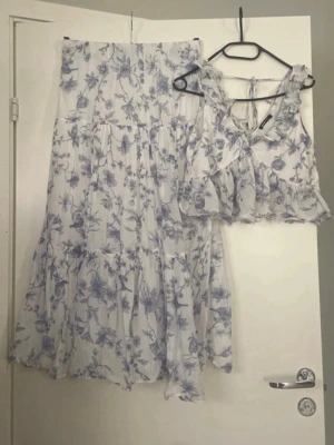 Vit långkjol med blå blommor och tillhörande top - Supersöt långkjol i vitt med blått blommönster och volangdetaljer. Kjolen har en luftig och flowy passform, perfekt för varma dagar. Matchande topp med volang och knyt i ryggen finns också. Materialet känns lätt och somrigt, troligen polyester eller bomull. Kjol storlek M o top strl S men den passar m också