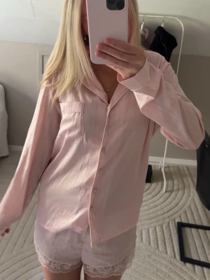 Ljusrosa pyjamas med spetsdetaljer - Söt ljusrosa pyjamas med långärmad skjorta och matchande shorts. Skjortan har knappar framtill och en bröstficka, medan shortsen har fina spetsdetaljer längs bensluten. Mjukt och lätt material som känns skönt mot huden. Helt oanvänt.
