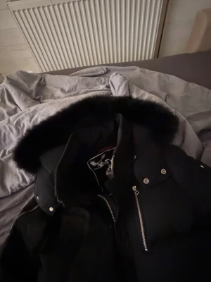 Mose knuckles jacka äkta  - Säljer en svart pufferjacka från Moose Knuckles med stor huva och svart fuskpäls. Jackan har silvriga dragkedjor, tryckknappar och två stora fickor framtill. Perfekt för kalla vinterdagar och riktigt snygg streetstil. Storlek M/S