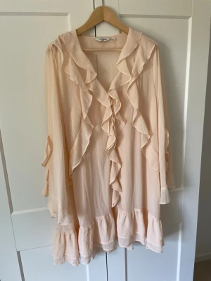 Ljus aprikos volangblus med långa ärmar - Romantisk blus med volangdetaljer längs fram- och nederkant. Blusen har lång ärm och är tillverkad i ett tunt, flowy material som ger ett luftigt intryck. Perfekt för dig som gillar en feminin och somrig  stil.