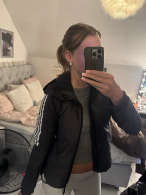 Svart Adidas vindjacka med huva - Snygg svart vindjacka från Adidas med klassiska vita ränder längs ärmarna och huva. Jackan har dragkedja framtill och Adidas-logga på sidan. Perfekt för en sportig och avslappnad stil. Materialet är lätt och vindtätt. Den är lite svart-grå. 