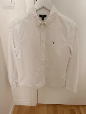 Vit skjorta från Gant med broderad logga - Klassisk vit skjorta från Gant med broderad logga på bröstet. Skjortan har lång ärm, knappar framtill och en stilren krage. Tillverkad i bomull och passar perfekt till en clean och tidlös stil.