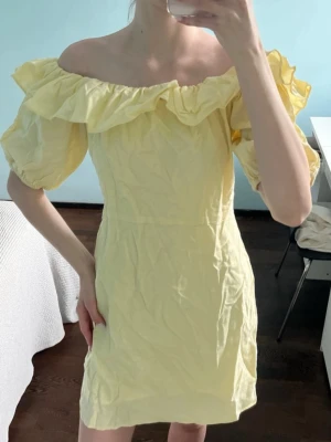Gul offshoulder klänning med volang - Söt gul kortklänning med offshoulder-design och puffiga korta ärmar. Klänningen har en bred volang runt axlarna och är tillverkad i ett lätt tyg, perfekt för varma dagar. Enkel och trendig modell som ger en romantisk vibe.