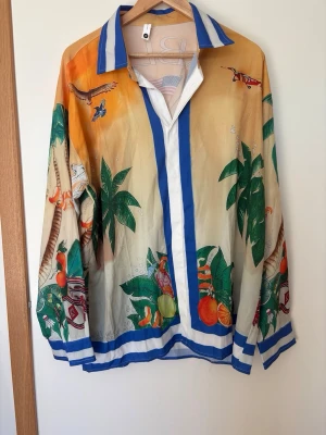Färgglad skjorta med palmmotiv - Unik skjorta med tropiskt motiv, palmer, frukter och djur i starka färger som orange, blå, grön och vit. Skjortan har blå och vita ränder på krage och kanter samt lång ärm. Perfekt för dig som gillar statement-plagg och färg.