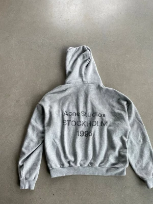 Grå hoodie Acne Studios  - Fet grå hoodie från Acne Studios med tryck på ryggen där det står 'Acne Studios STOCKHOLM 1996'. Matrial är heavy cotton med distressed details på luvan. Perfekt för dig som gillar streetwear eller något liknande. Påse ingår.