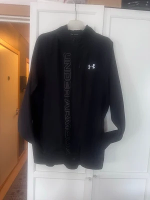 Svart Under Armour windbreaker - Snygg svart windbreaker från Under Armour med logga på bröstet och stort tryck längs dragkedjan. Tillverkad i ett mjukt och funktionellt material, perfekt för träning eller chill. Huva och fickor på sidorna ger extra komfort.