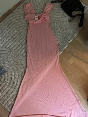 Lång rosa klänning med breda axelband - Säljer en elegant långklänning i mjukt rosa tyg med breda axelband och draperad urringning. Klänningen har en figurnära passform och är perfekt för speciella tillfällen. Materialet känns lent och följsamt mot kroppen. Helt ny, köpt från Cider. 