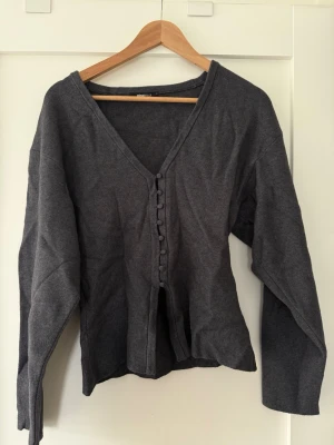 Mörkgrå kofta från Gina Tricot - Mörkgrå kofta från Gina Tricot med v-ringning och knappar framtill. Långärmad och i mjukt, tunt material som passar perfekt till lager-på-lager. Enkel och stilren design som funkar till många outfits.