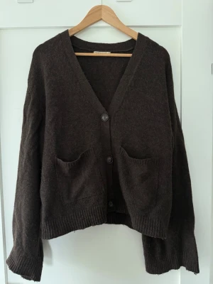 Brun kofta från Gina Tricot - Mysig brun kofta från Gina Tricot med v-ringning, tre knappar framtill och två stora fickor. Koftan har en avslappnad passform och ribbade muddar vid ärmslut och nederkant. Perfekt för lager på lager och enkel att styla till olika outfits.