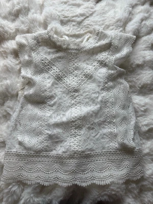 Vit spetsblus med hög krage - Supersöt vit blus i spets med hög krage och fina spetsdetaljer. Blusen är ärmlös och har ett vackert mönster samt vågig kant nertill. Perfekt för dig som gillar romantisk och feminin stil.