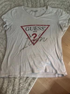 Vit t-shirt med logga från Guess - Vit t-shirt från Guess med ikonisk röd trekantig logga och glittrig silvertext på bröstet. T-shirten har korta ärmar och v-ringning. Perfekt för dig som gillar klassiska märken med en twist.