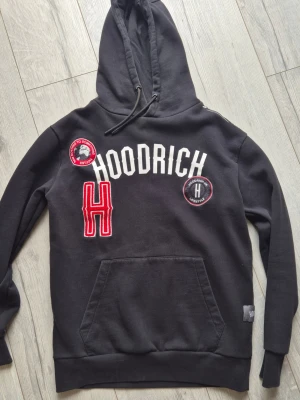 Svart hoodie från Hoodrich - Svart hoodie från Hoodrich med stor vit logga på bröstet, röda och svarta patchar samt en stor röd broderad H. Klassisk känguruficka framtill och justerbar huva med snören. Mjuk bomullskänsla och ribbade muddar vid ärmar och nederkant.