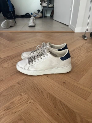 Vita sneakers från Crime london - Snygga vita sneakers från Crime London med blå detalj på hälen och vita snören. Skorna har en klassisk låg profil och är tillverkade i läder med en robust gummisula. Perfekta för en clean och stilren look.
