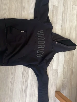 Svart hoodie från Hoodrich - Säljer en svart hoodie från Hoodrich med stor logga på bröstet. Hoodien har huva med knappdetaljer och en stor magficka. Tillverkad i mjukt material som känns skönt mot huden. Perfekt för dig som gillar streetwear och vill ha en clean look.
