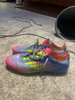 F50  - Säljer ett par Adidas X Speedflow fotbollsskor i blått, rosa och gult. Skorna har snörning, låg profil och är tillverkade i syntetmaterial för lätt vikt och bra bollkontroll. Perfekta för snabba spelare som vill sticka ut på planen.