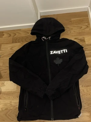 Svart vindjacka från Zavetti - Snygg svart vindjacka från Zavetti med huva och dragkedja framtill. Jackan har reflekterande logga på bröstet och ett stilrent lönnlövsmärke. Två fickor med dragkedja och justerbar huva. Perfekt för blåsiga dagar och streetwear-stil.