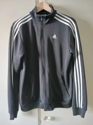 Svart Adidas track jacket Storlek L - Klassisk svart Adidas track jacket med vita ränder längs ärmarna och dragkedja framtill. Jackan har hög krage, två fickor med dragkedja och Adidas-logga på bröstet. Tillverkad i mjukt och bekvämt material, perfekt för träning eller chill. Storlek L