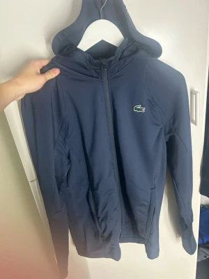 Mörkblå vindjacka från Lacoste - Snygg mörkblå vindjacka från Lacoste med dragkedja framtill och huva. Jackan har det klassiska krokodilmärket på bröstet och diskret tryck på ryggen. Tillverkad i ett lätt och vindtätt material, perfekt för blåsiga dagar. Två fickor framtill och smal passform.