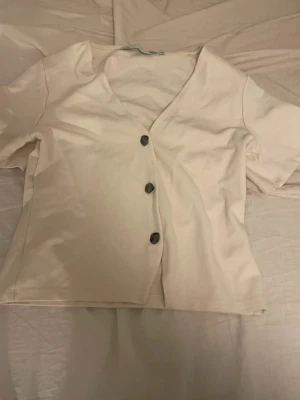 Vit v-ringad topp med knappar - Snygg vit topp med v-ringning och tre dekorativa knappar framtill. Kortärmad och i mjukt bomullsmaterial, perfekt för en avslappnad och stilren look.
