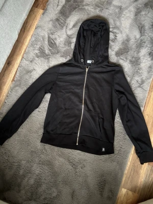 Svart hoodie med dragkedja från FBSISR - Säljer en svart hoodie från FBSISR i storlek M. Hoodien har dragkedja framtill, två fickor och en klassisk huva med metallöljetter. Enkel och stilren design som passar till det mesta.
