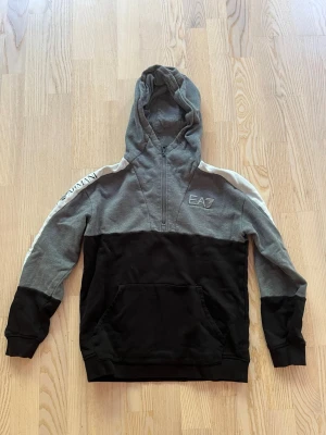 Grå och svart hoodie från EA7 - Snygg hoodie från EA7 Emporio Armani i grått och svart med vit detalj längs axlarna. Halvlång dragkedja. Storlek 160. 
