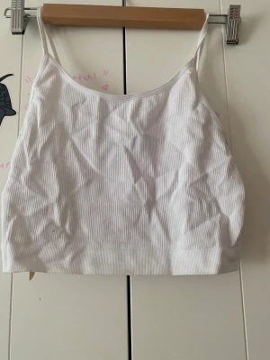 Vit ribbad linnetopp - Säljer en enkel och stilren vit ribbad linnetopp med smala axelband. Toppen är croppad och har en mjuk, stretchig känsla som passar perfekt till jeans eller kjol. Perfekt för varma dagar eller som lager under en skjorta.