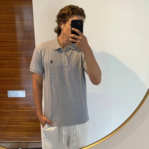 Grå pikétröja från Ralph Lauren - Klassisk grå pikétröja från Ralph Lauren med den ikoniska broderade loggan på bröstet. Tröjan har korta ärmar, knappar vid halsen och är tillverkad i mjuk bomull. Perfekt för en clean och avslappnad stil. Storlek M, jag är 176 cm.